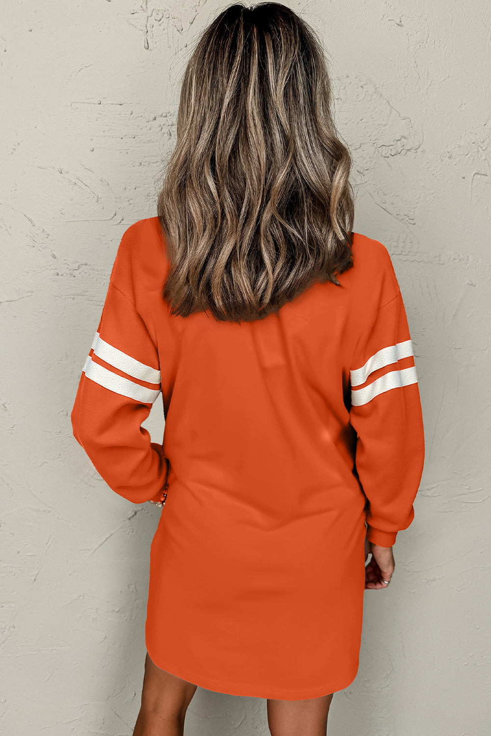 Back view of a model in an orange colorblock athletic dress showcasing long sleeves and sporty stripes, with flowing hair.