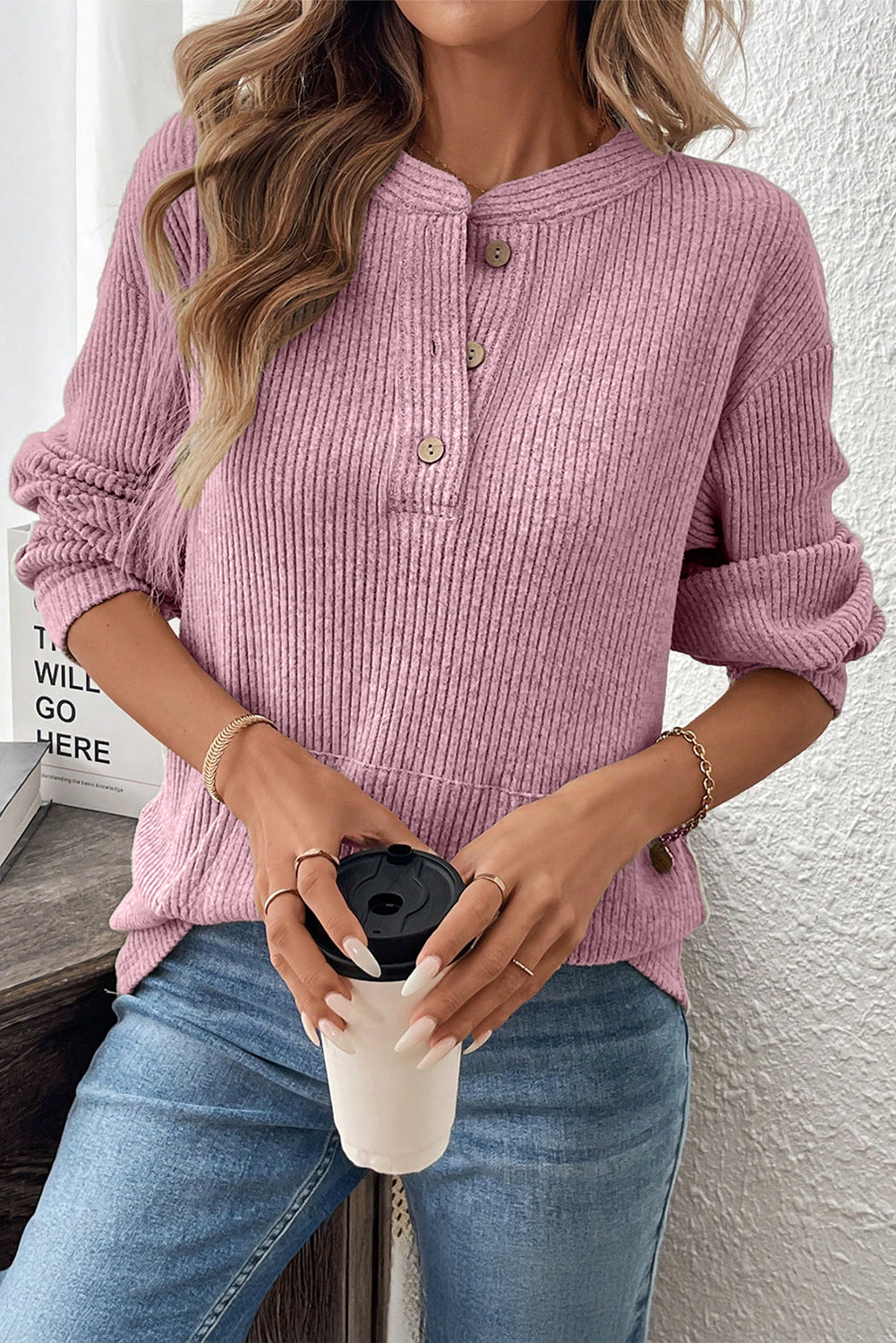 Person wearing a pink ribbed sweater and blue jeans, holding a white coffee cup with a black lid.