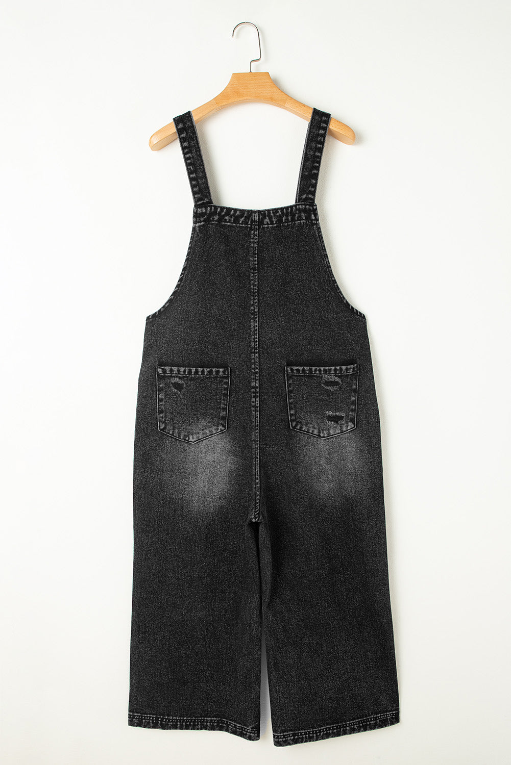 Black denim overalls hanging on a wooden hanger against a white background