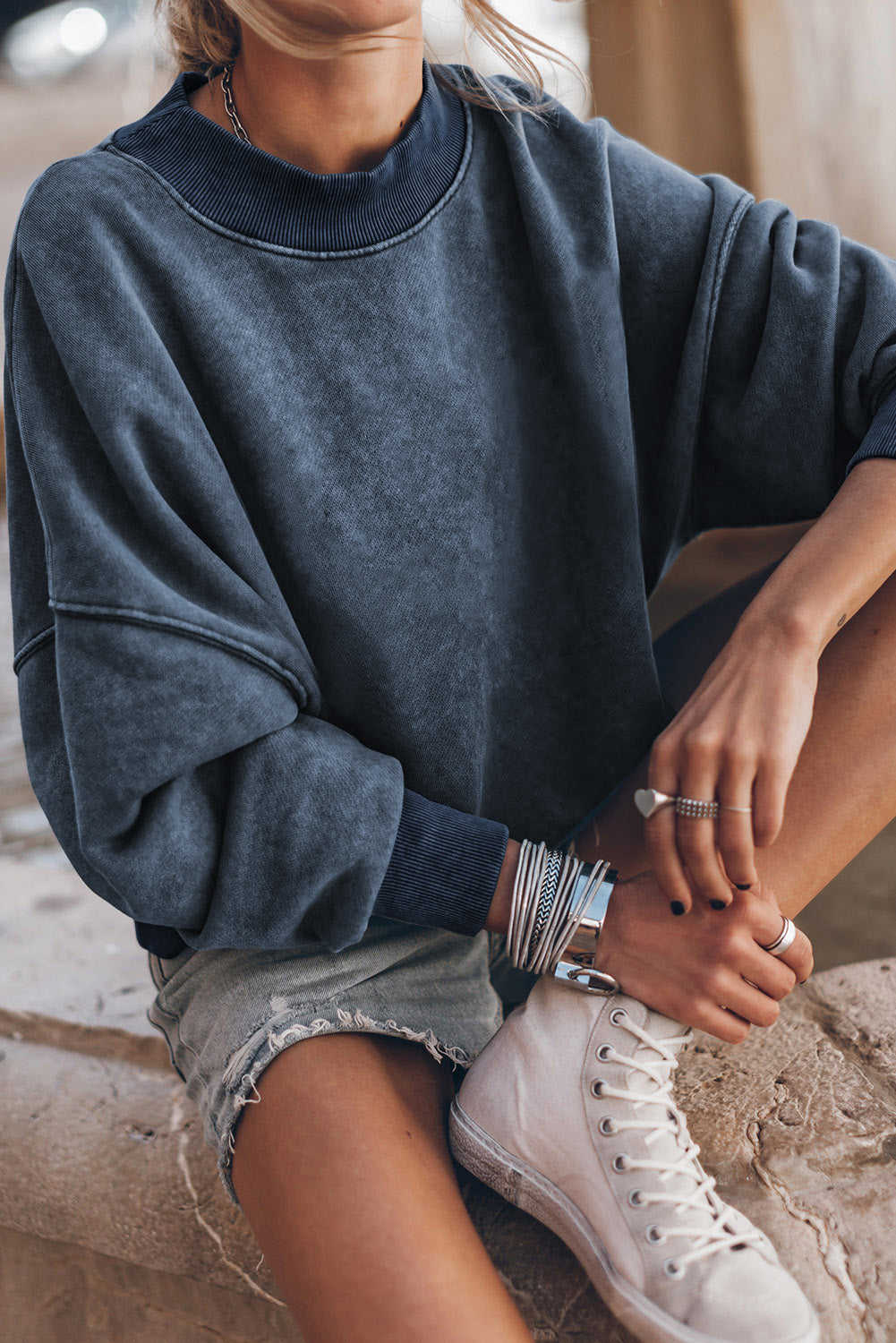 Person wearing a blue sweatshirt and white sneakers sitting on a stone surface.