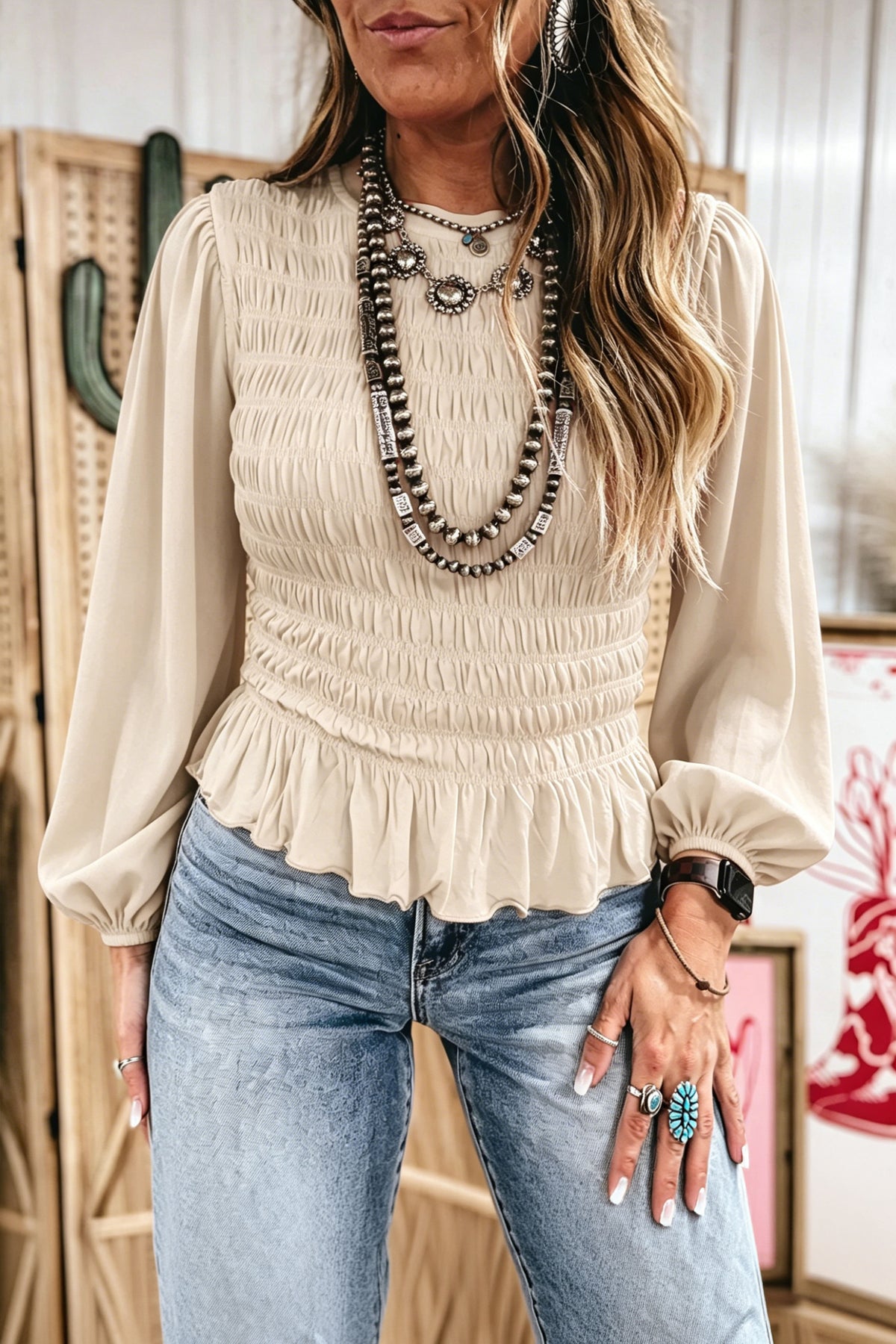 A woman wearing a Chic jet stream Smocked Ruffle Hem Boho Blouse with a ruffled hem, paired with light-wash blue jeans and layered statement necklaces, standing in a styled indoor setting.