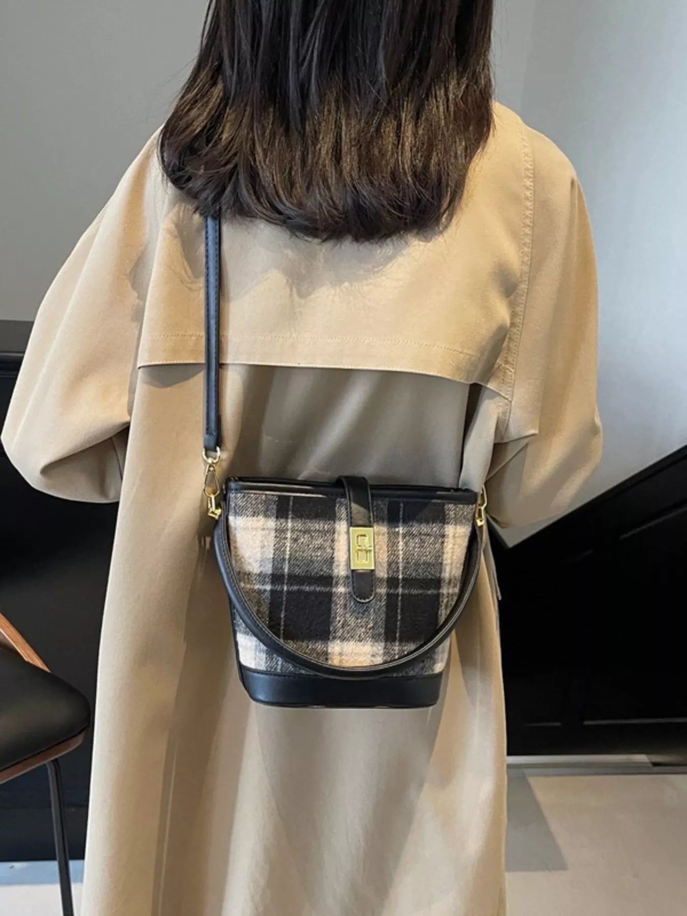 Person wearing a beige trench coat with a plaid handbag over their shoulder indoors.
