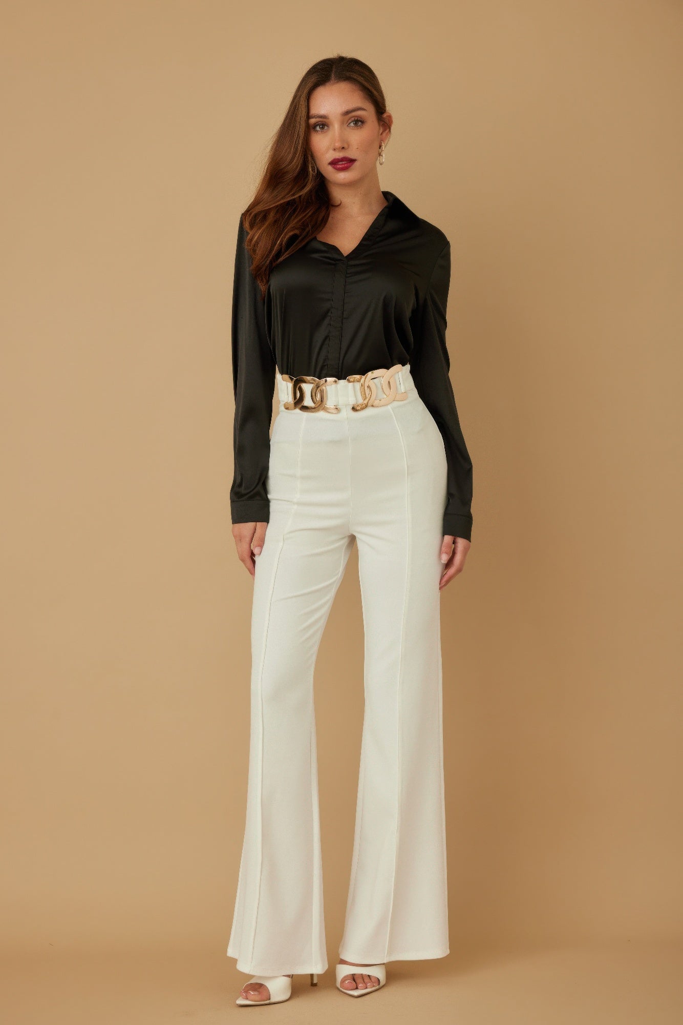 Woman wearing a black top and white pants with a gold belt against a beige background
