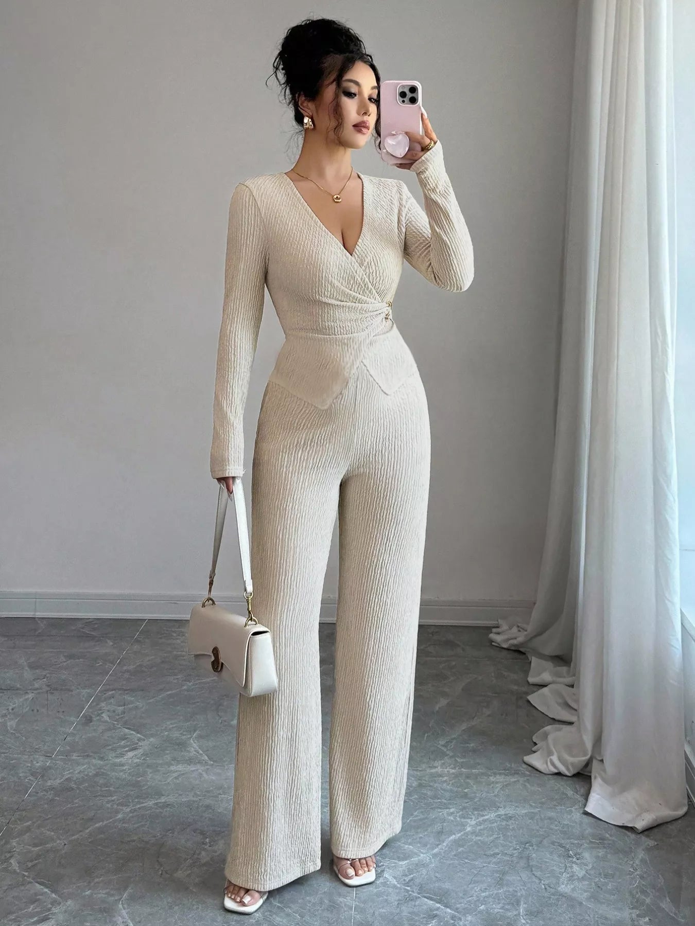 Woman in a beige jumpsuit taking a mirror selfie in a room with light gray walls and white curtains.
