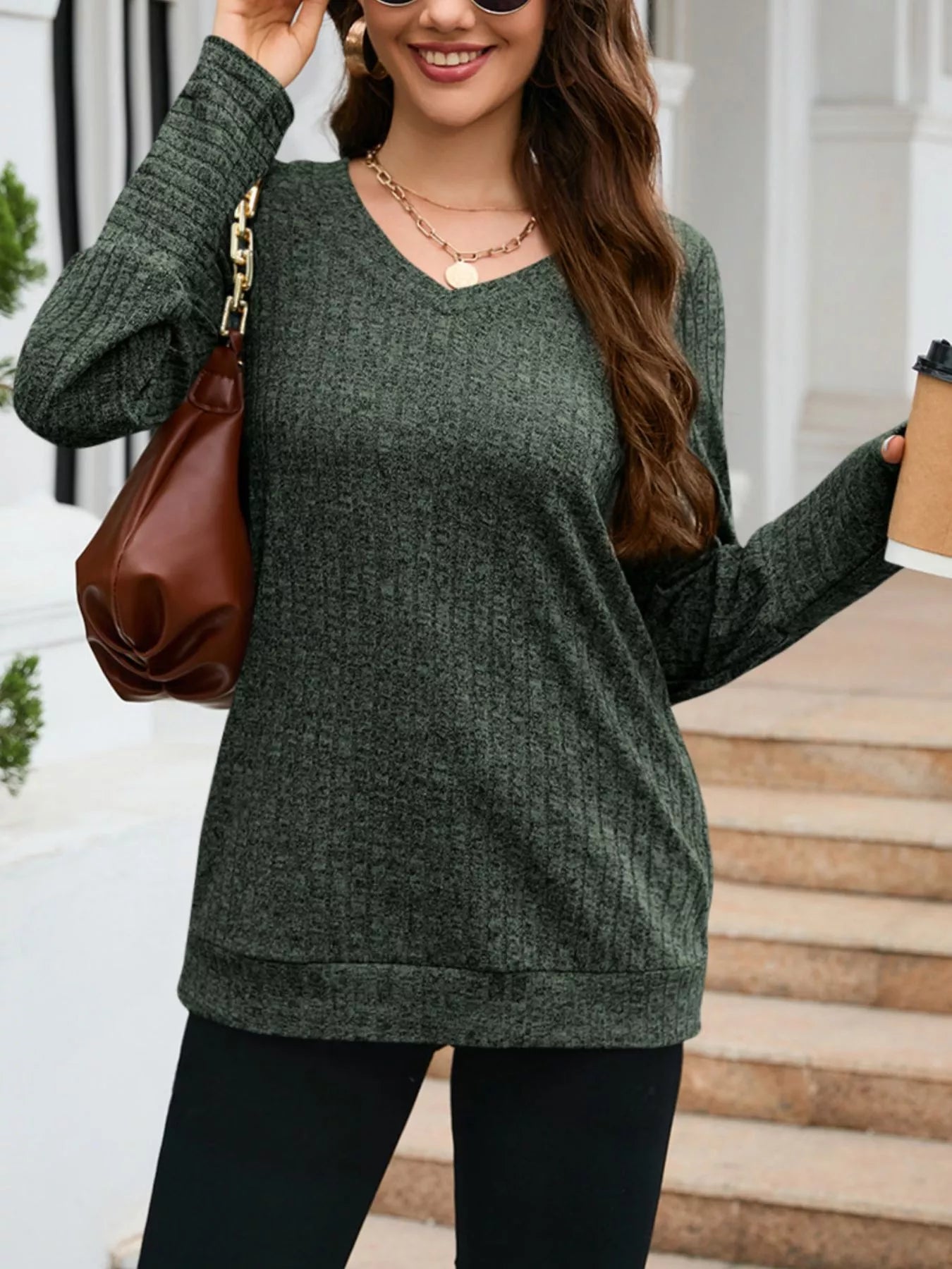 Woman wearing a green sweater holding a brown bag and a coffee cup outdoors.