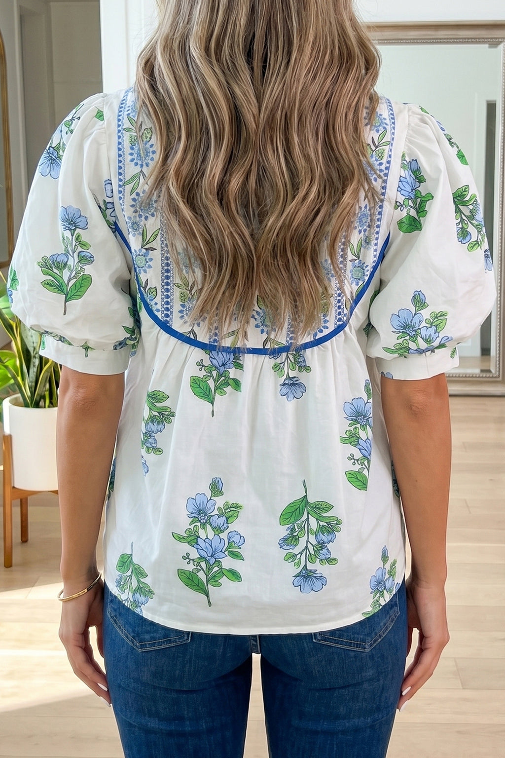 Person wearing a white blouse with floral embroidery in a room with plants and furniture. Back view