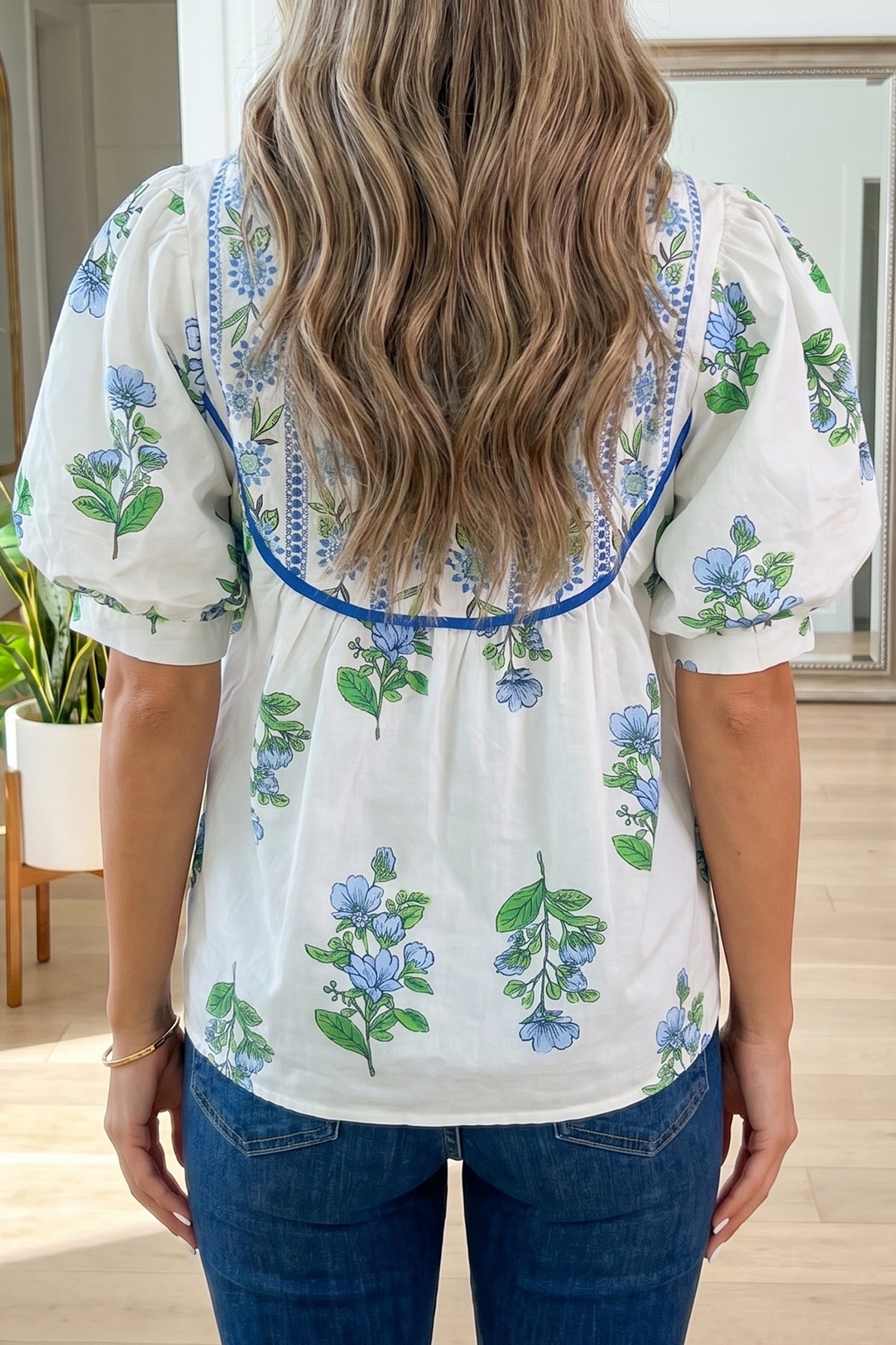Person wearing a white blouse with floral embroidery in a room with plants and furniture. Back view