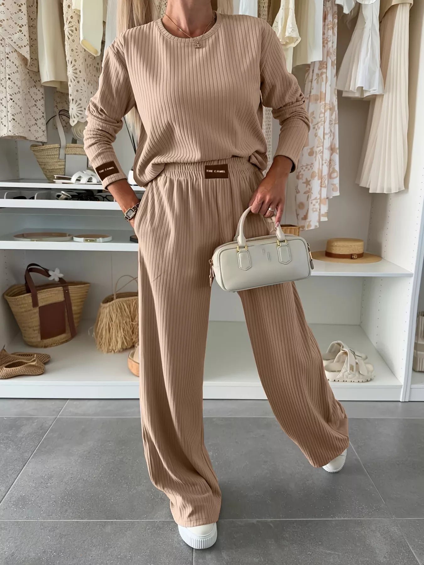 Person wearing a beige ribbed outfit holding a white handbag in a store setting.
