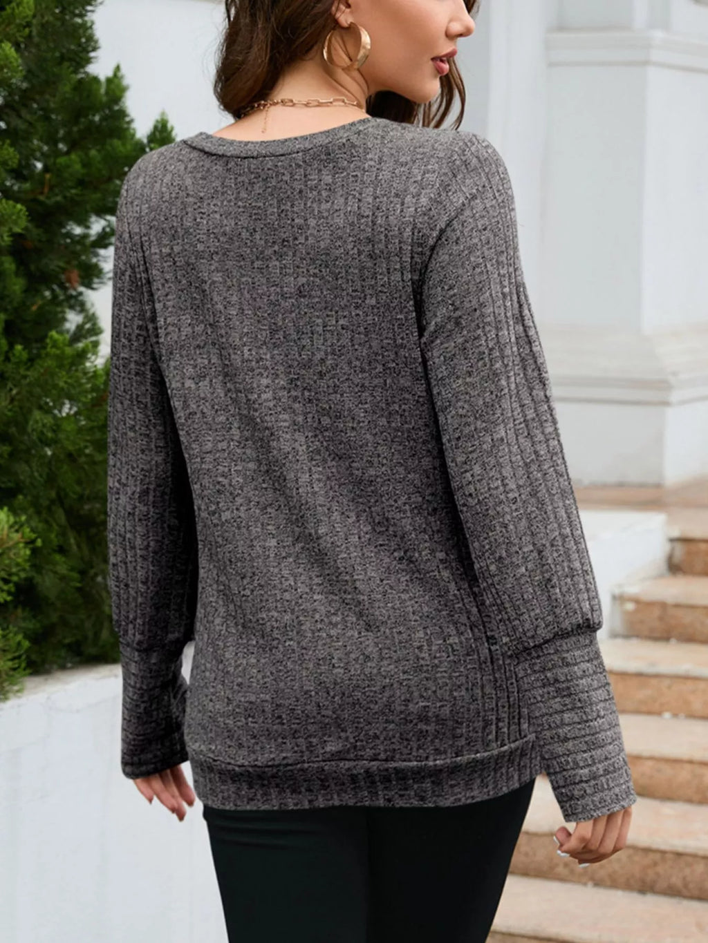 Woman wearing a gray sweater standing outdoors with steps and greenery in the background
