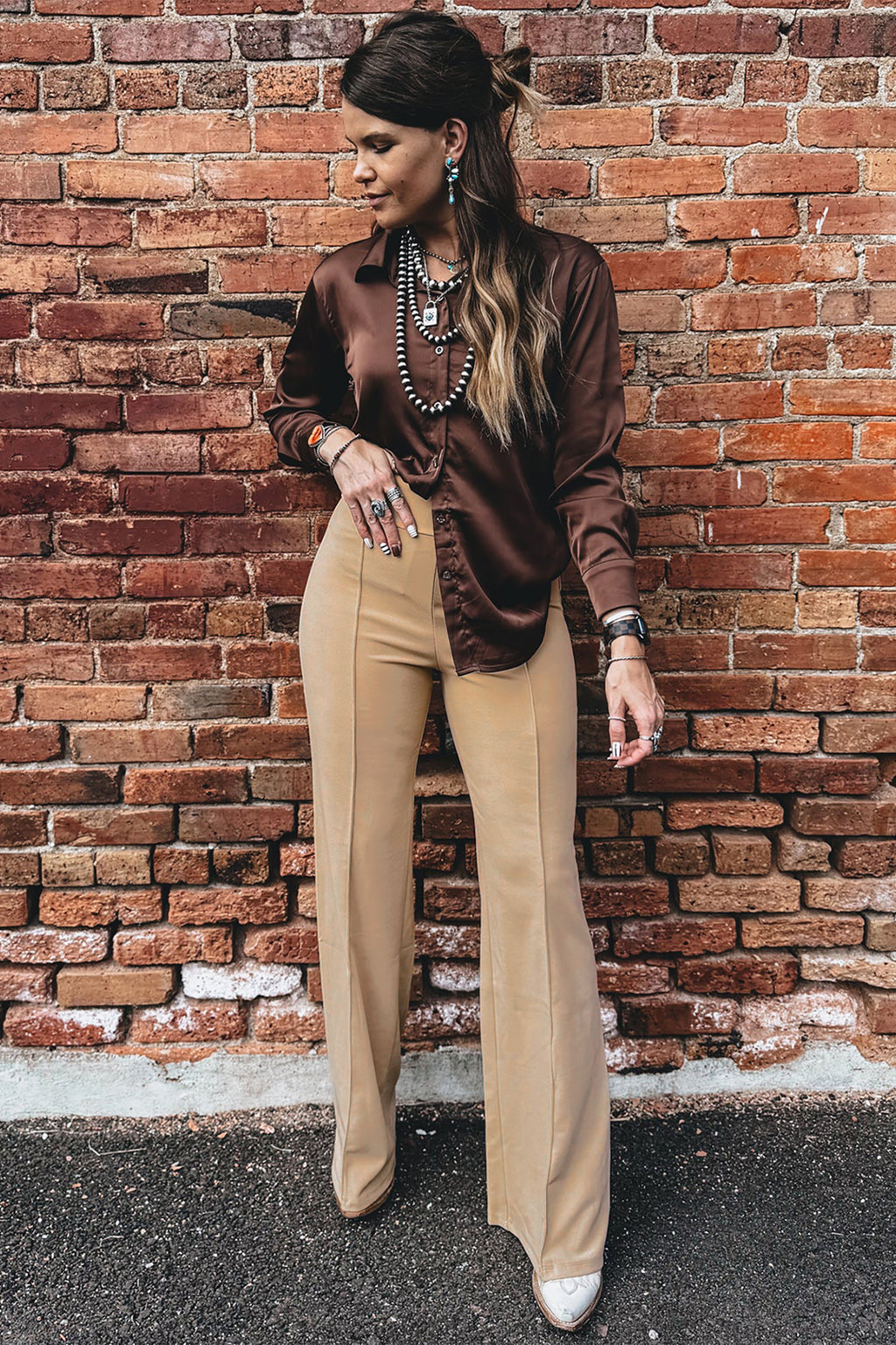 A model stands against a red brick wall wearing a dark brown satin long-sleeve blouse tucked into Stylish women's High-Waist Parchment Pants, accessorized with layered necklaces and bracelets.