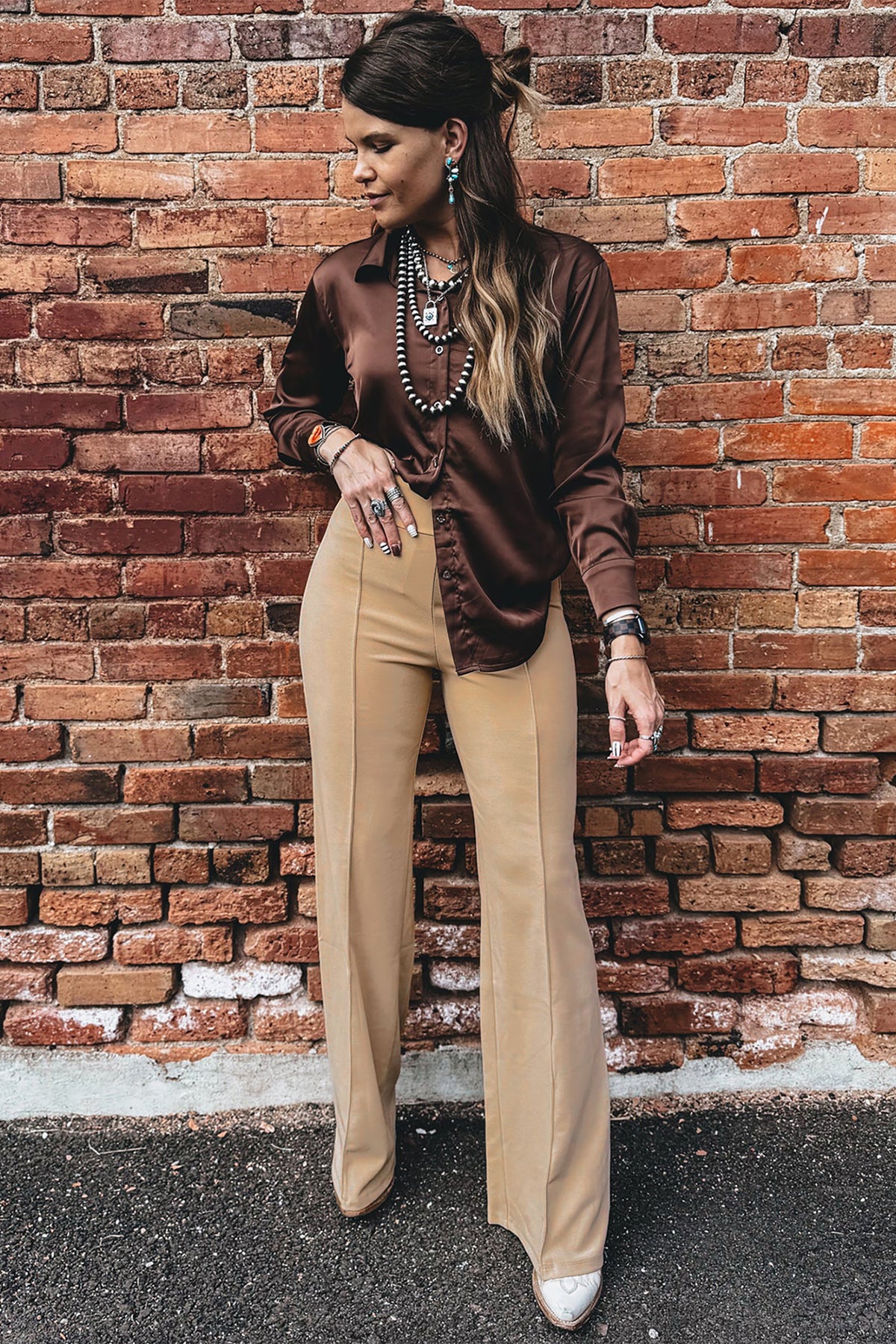 A model stands against a red brick wall wearing a dark brown satin long-sleeve blouse tucked into Stylish women's High-Waist Parchment Pants, accessorized with layered necklaces and bracelets.