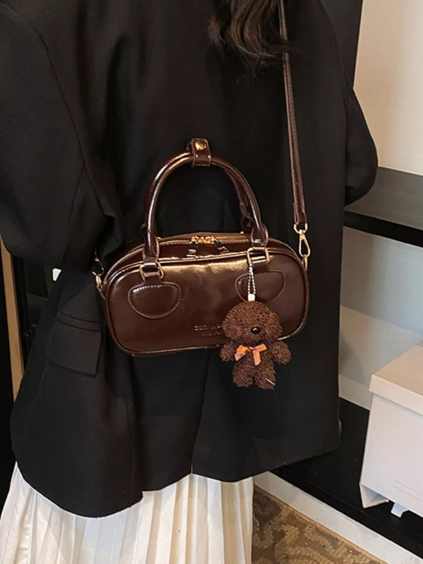 Brown handbag with a teddy bear charm held by a person wearing a black outfit.