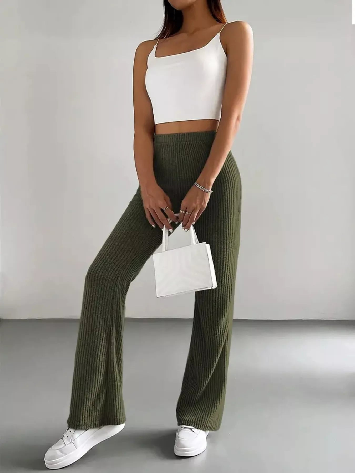 Person wearing a white top and green ribbed pants holding a small white handbag against a plain background