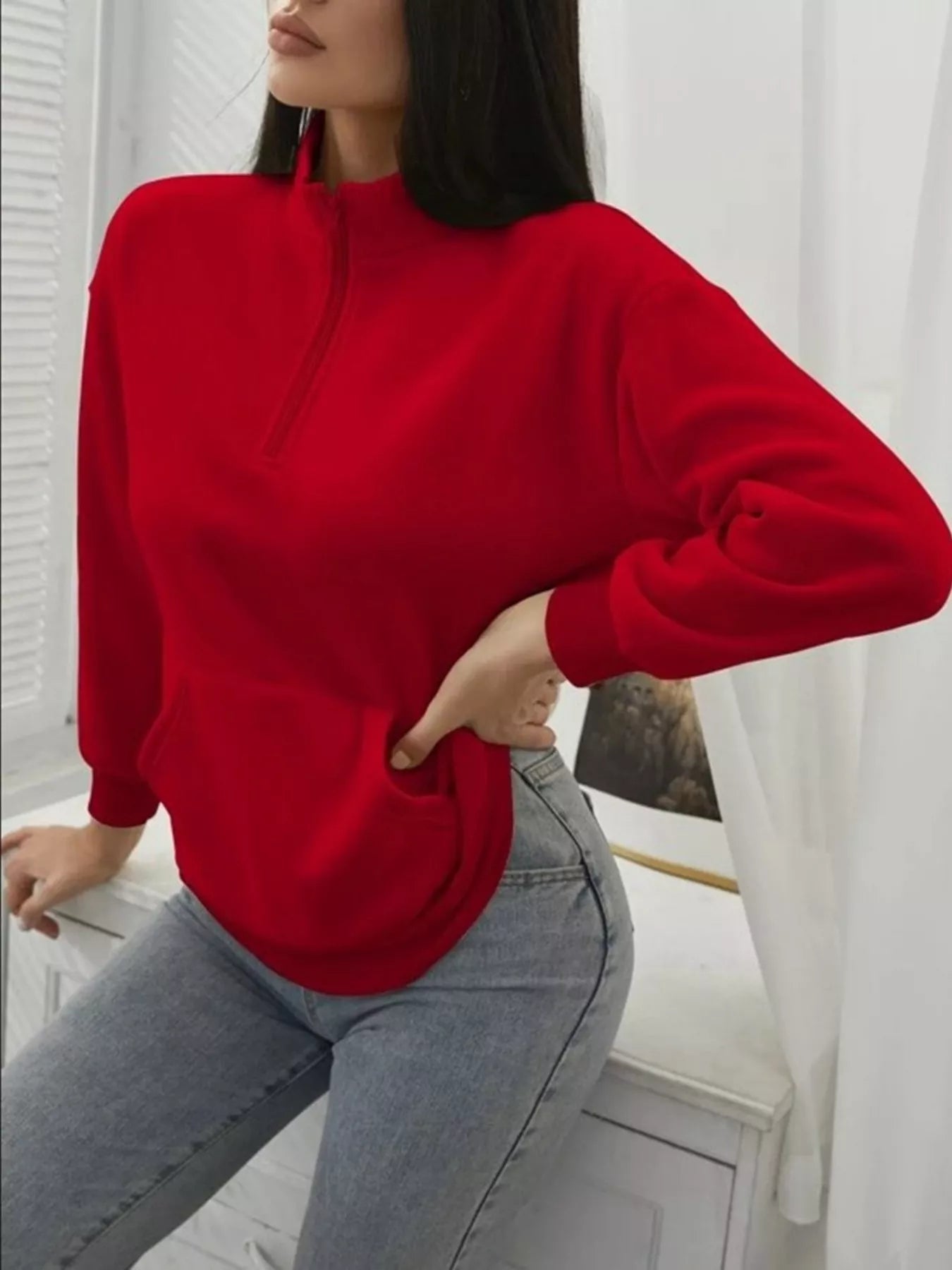 Person wearing a red sweater and gray jeans against a white curtain background
