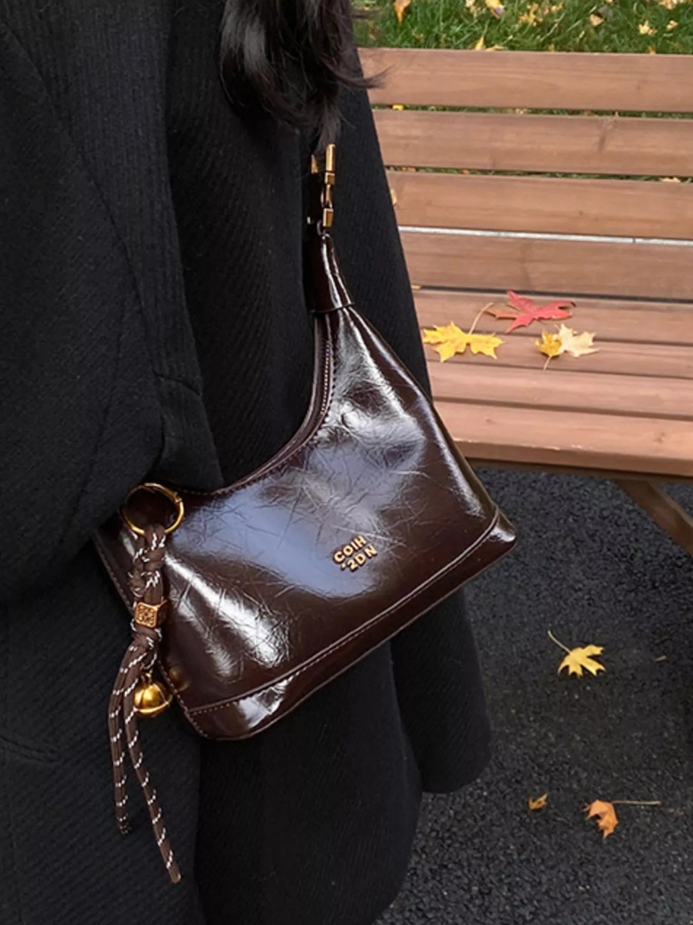 Brown leather handbag with gold accents held by a person in a black coat, with a wooden bench and autumn leaves in the background.