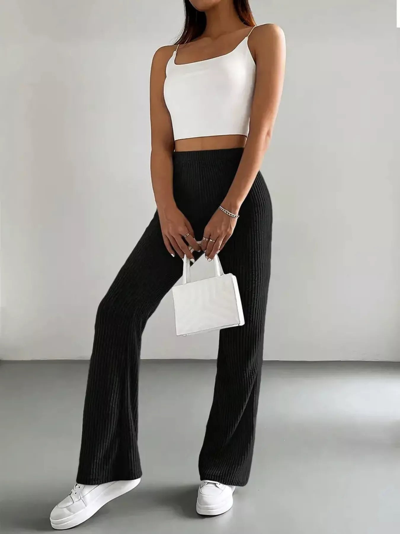 Person wearing a white crop top and black wide-leg pants, holding a small white handbag against a plain background.