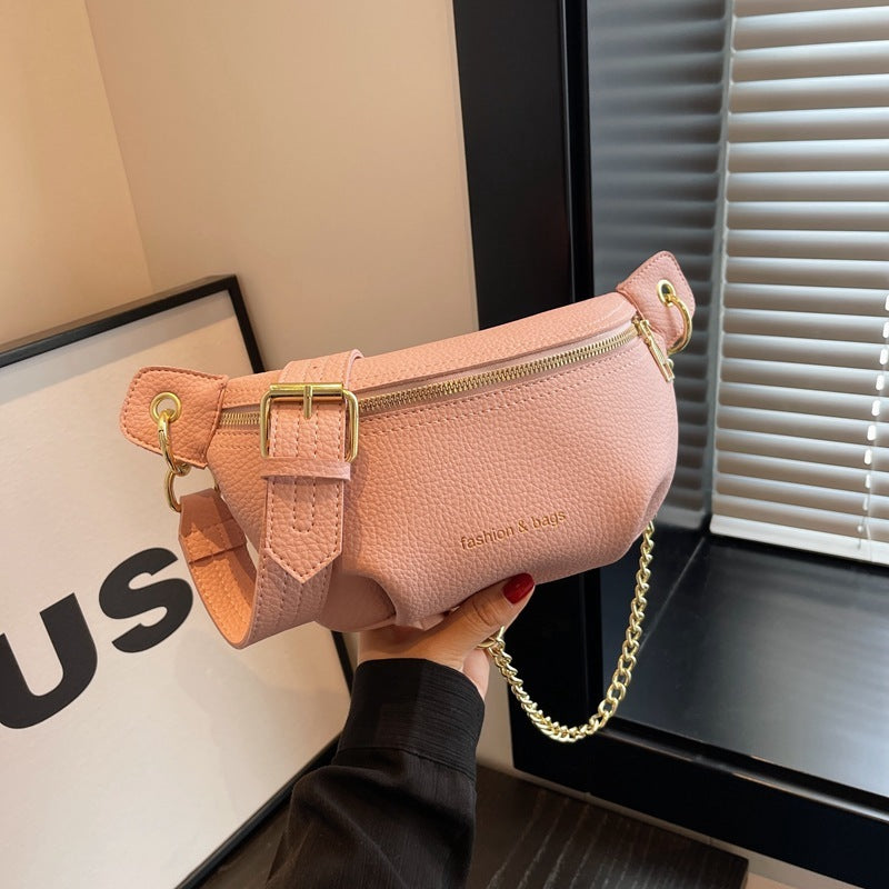 Pink waist bag with gold chain held by a hand, with a blurred indoor background.