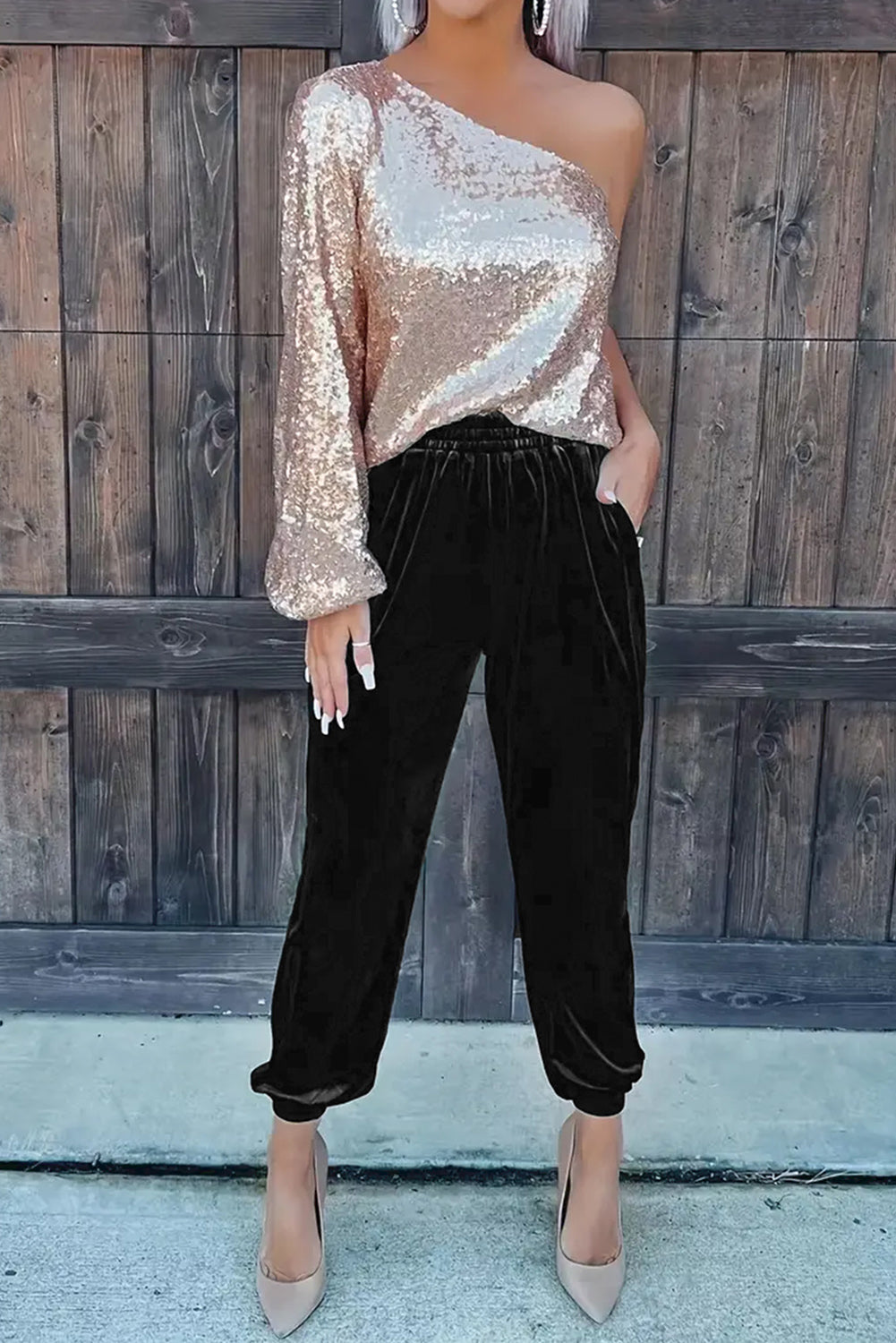 Person wearing a one-shoulder sequin top and black velvet pants against a wooden fence.