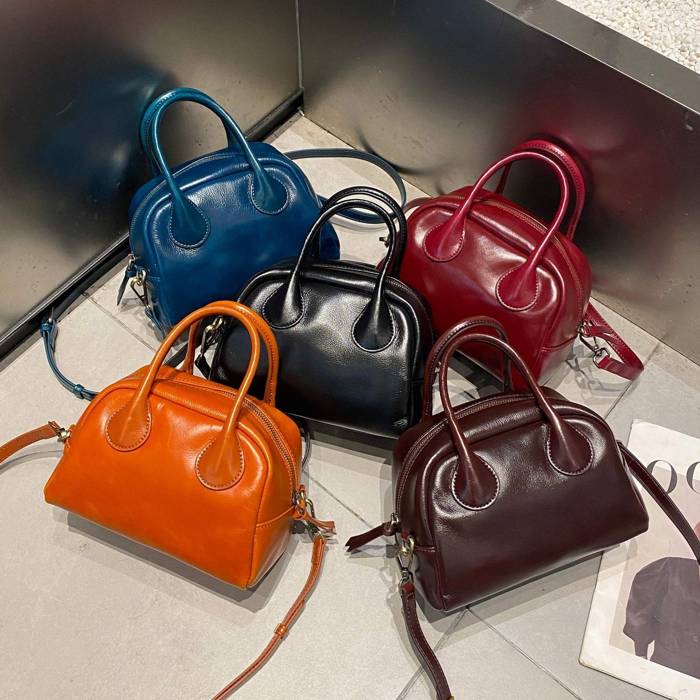 Five handbags in blue, black, red, orange, and brown on a reflective surface.