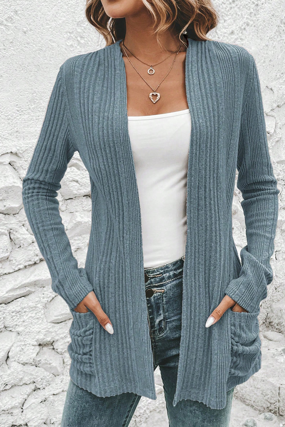 Person wearing a blue ribbed cardigan over a white top and jeans against a stone wall background