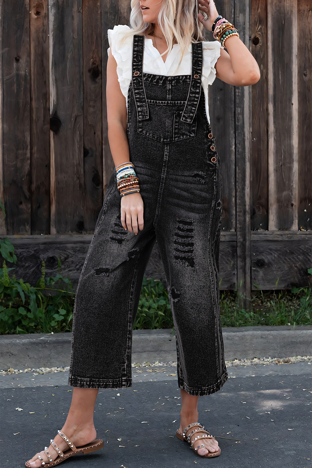 Woman wearing black distressed overalls with a white top against a wooden fence background
