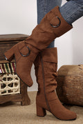 Brown knee-high boots with decorative buckles worn by a person, with a neutral background.