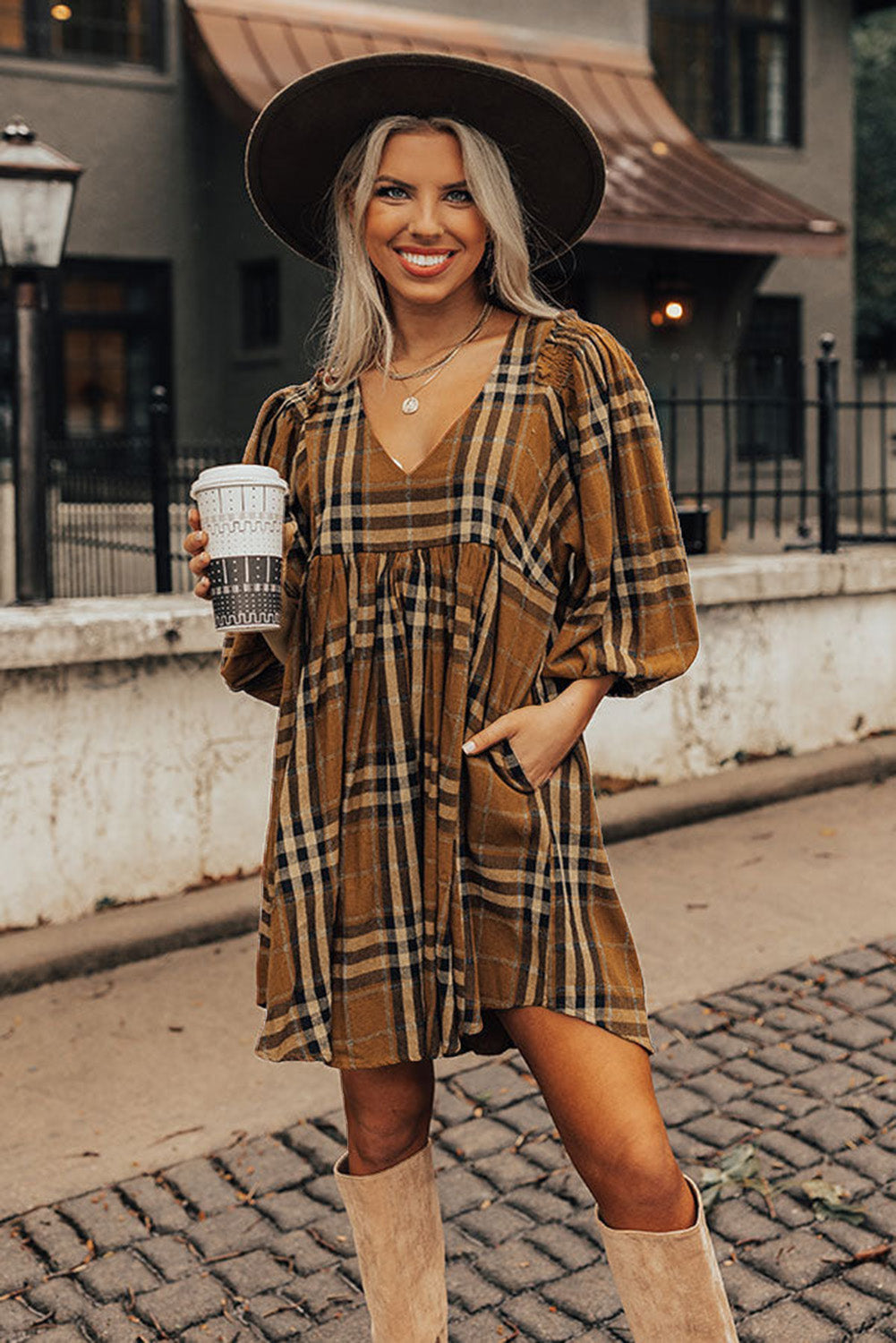 Woman in plaid dress and hat holding a coffee cup on a street.
