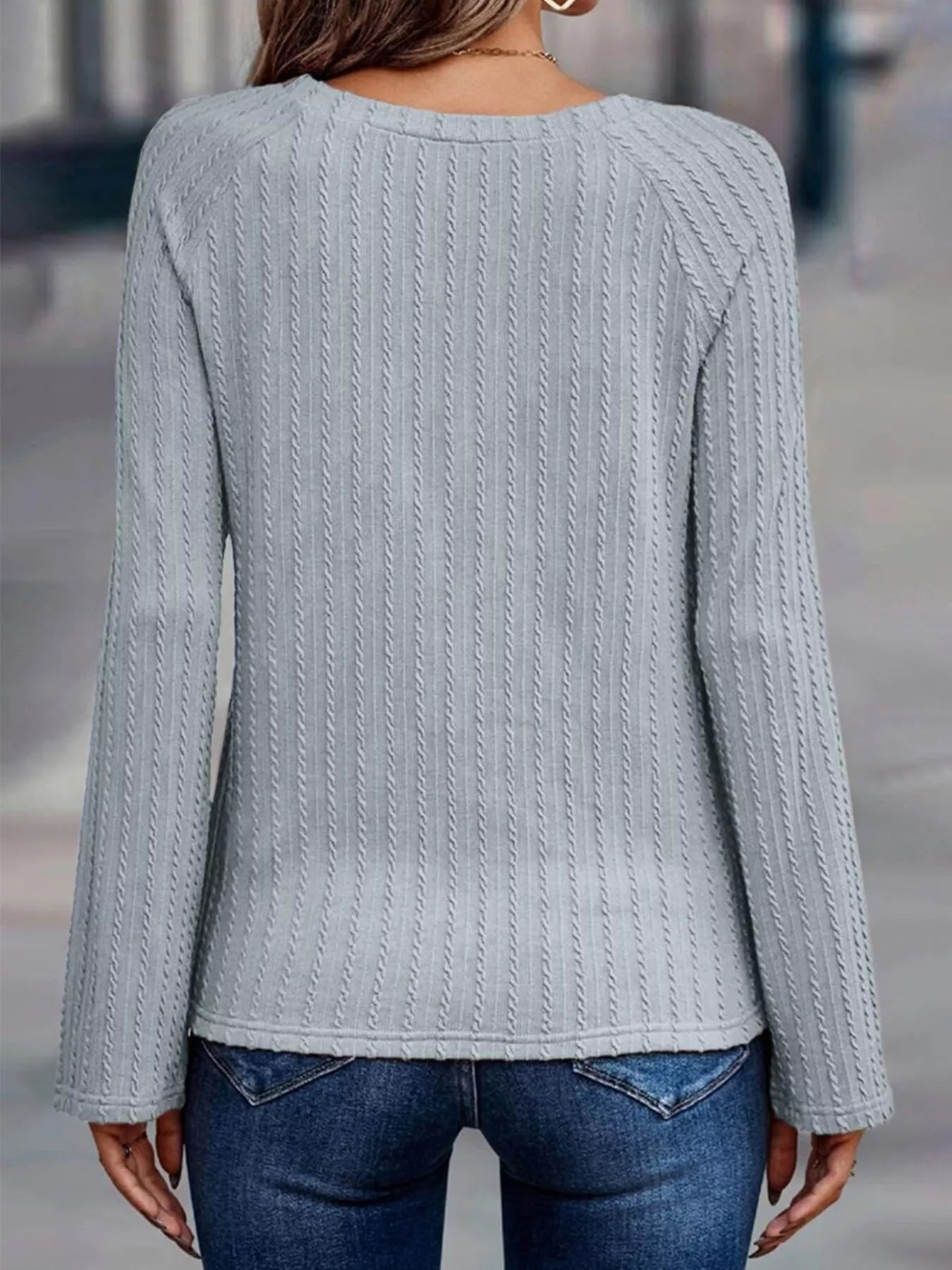 Person wearing a light gray cable knit sweater and blue jeans on a blurred street background