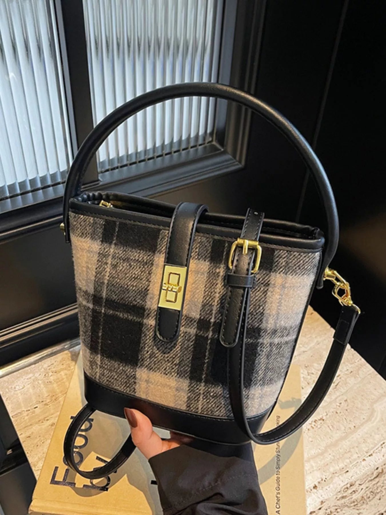 Plaid handbag with black handles and strap held by a person indoors.