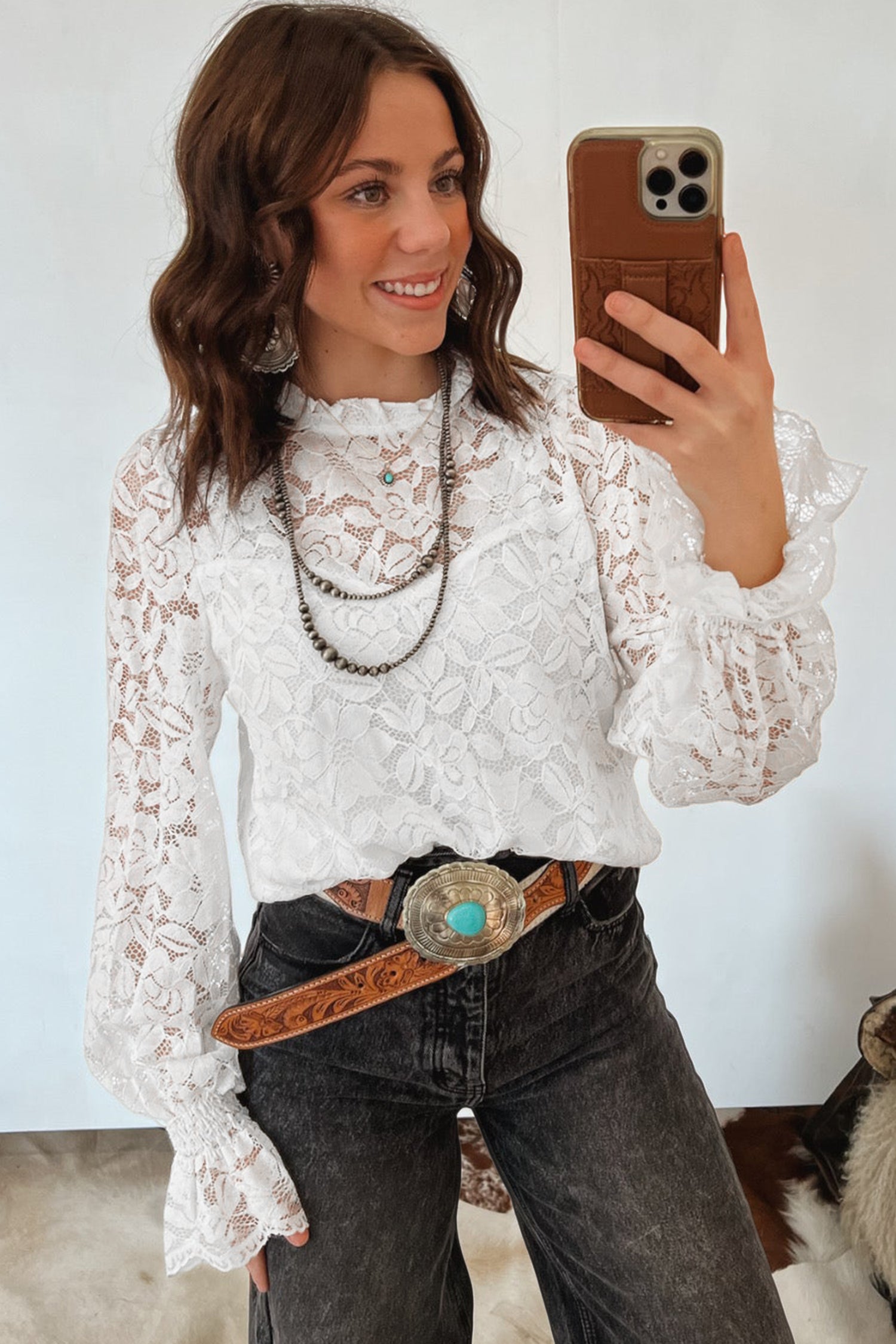 Woman taking a mirror selfie wearing a white lace top and dark jeans with a brown belt.