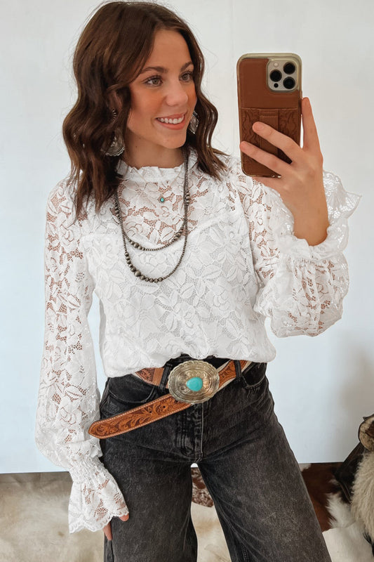 Woman taking a mirror selfie wearing a white lace top and dark jeans with a brown belt.