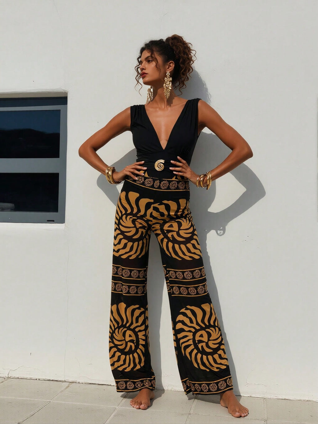 Woman wearing a black top and patterned pants standing against a white wall.
