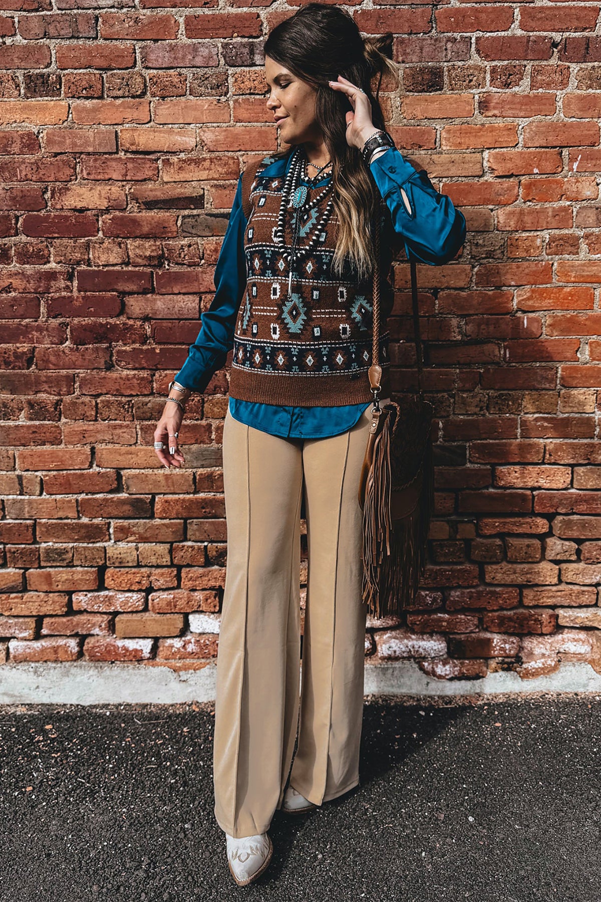 A model stands against a red brick wall wearing a bohemian-style outfit featuring a turquoise long-sleeve shirt, a brown patterned vest, Stylish women's High-Waist Parchment Pants, and a brown fringe crossbody bag.