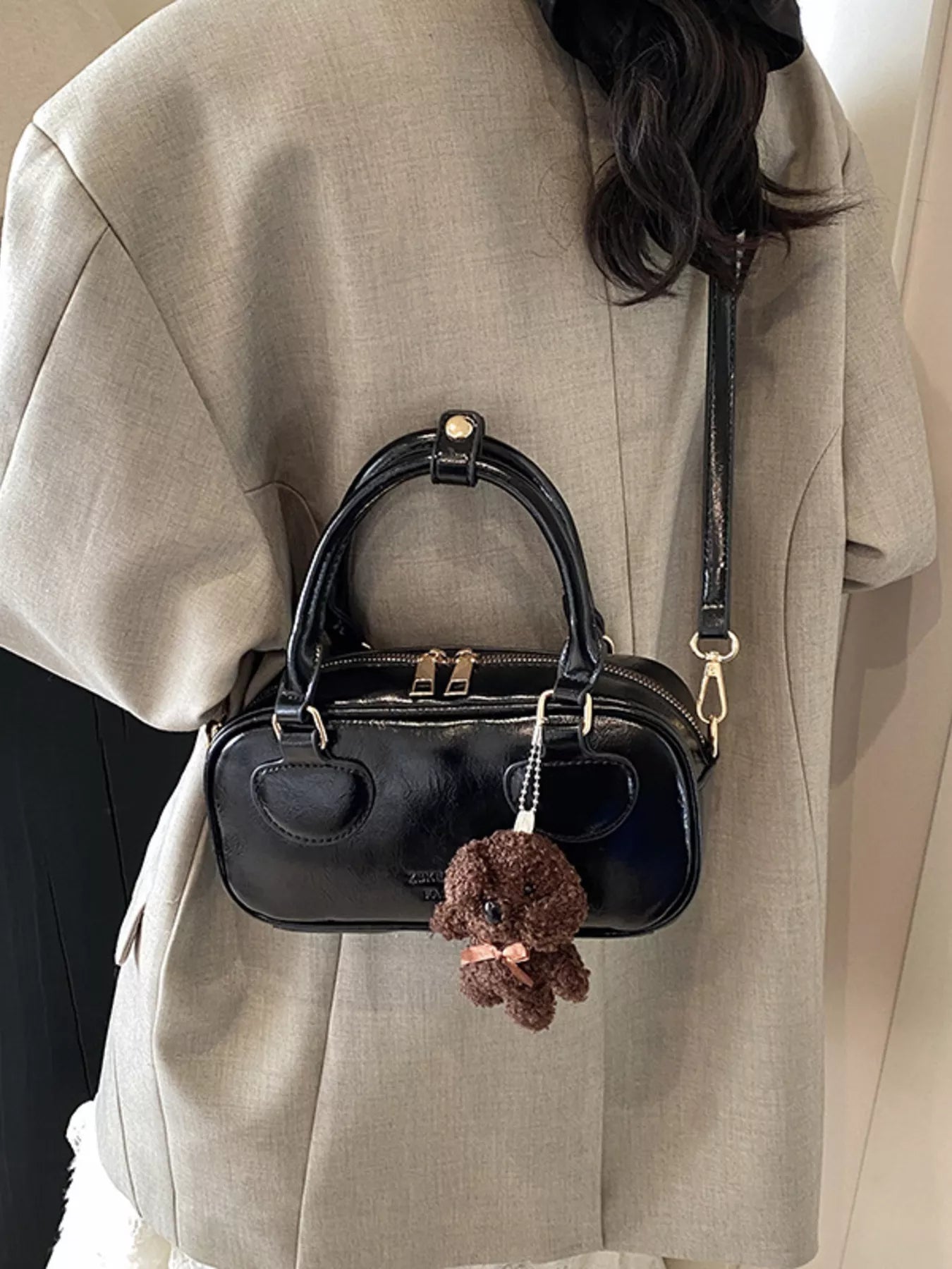 Black handbag with a small brown plush toy attached, worn by a person in a beige coat.