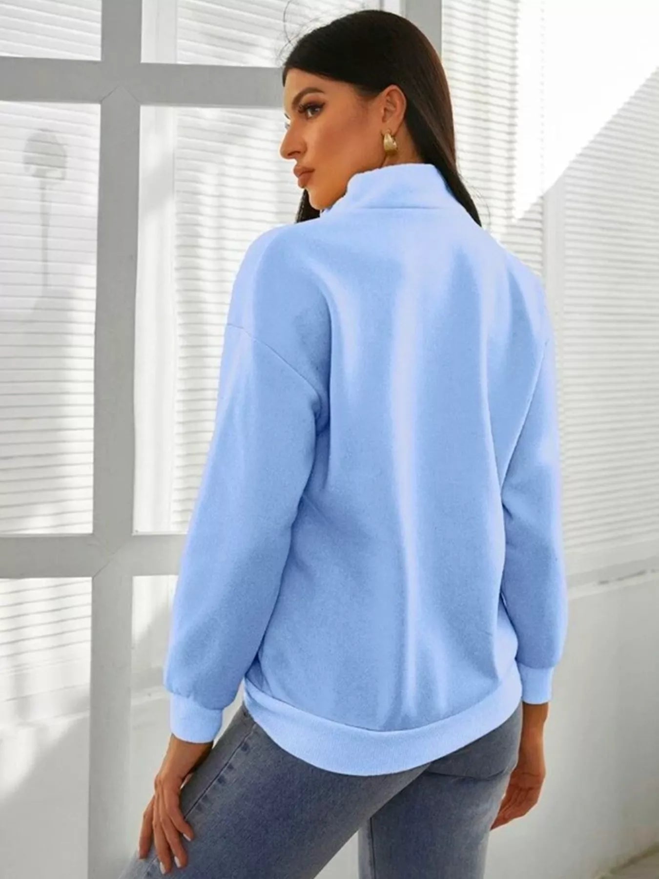 Woman wearing a light blue sweater in a room with white walls and windows.