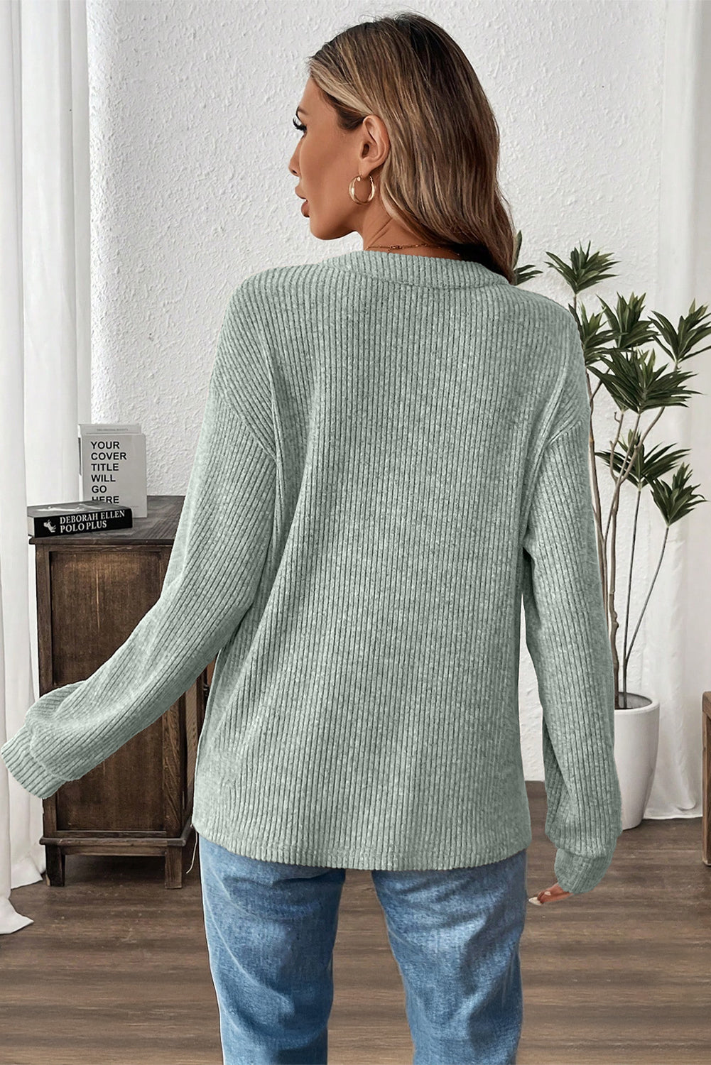 Woman wearing a green ribbed sweater and blue jeans in a room with a plant and wooden cabinet.