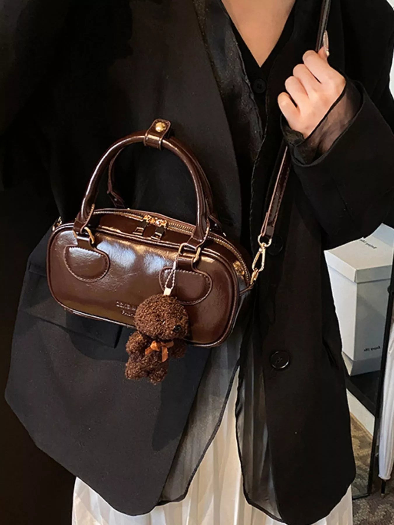 Brown handbag with a teddy bear keychain held by a person wearing a black outfit.
