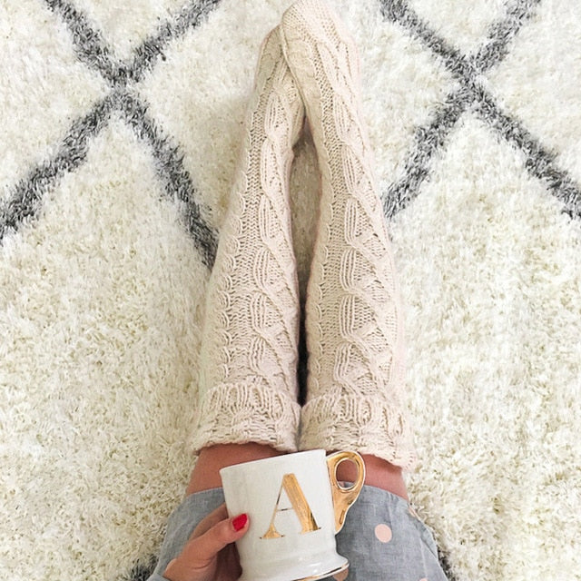 Person wearing a cozy beige sweater holding a mug on a soft surface with a checkered pattern.