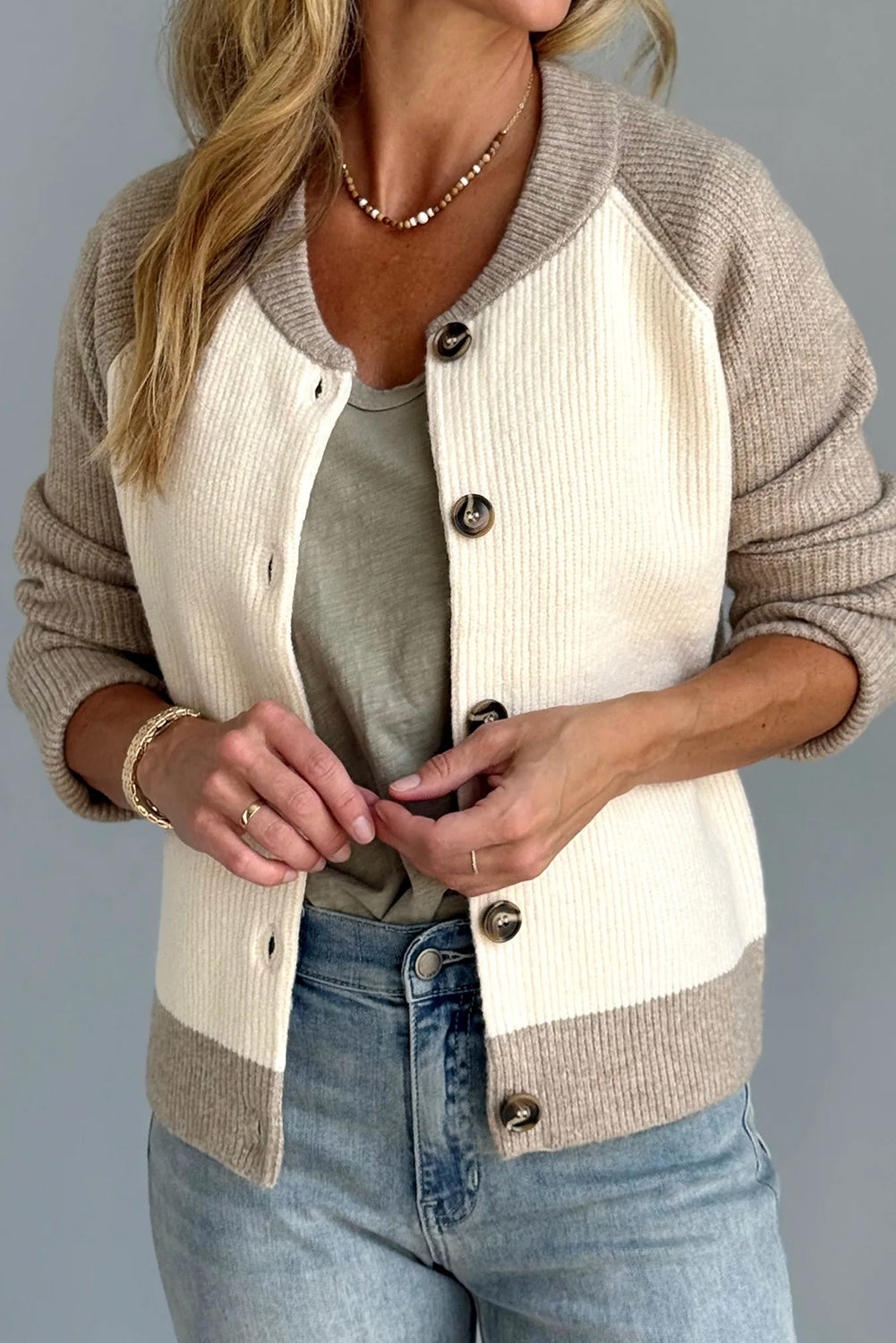Person wearing a beige and white cardigan over a gray shirt with blue jeans on a gray background