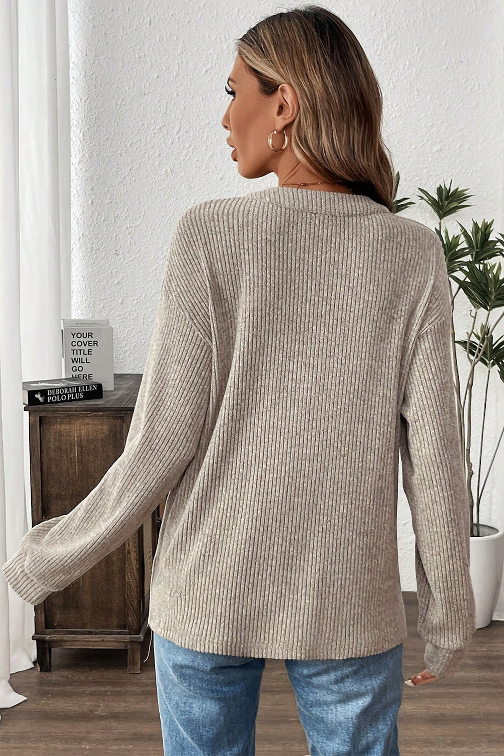 Woman wearing a beige ribbed sweater and blue jeans in a room with a wooden cabinet and plant.