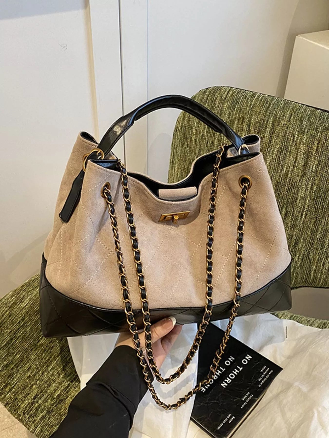 Beige handbag with black accents and gold chain held by a person on a green couch.