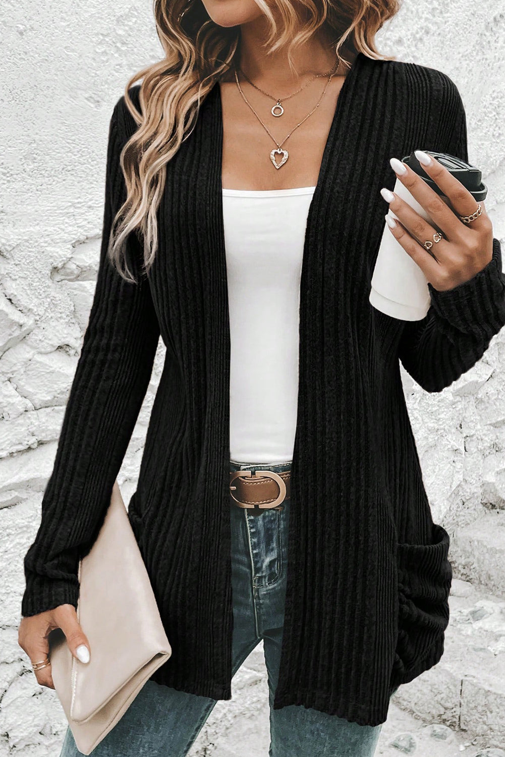 Person wearing a black cardigan over a white top and blue jeans, holding a beige clutch and a black cup.