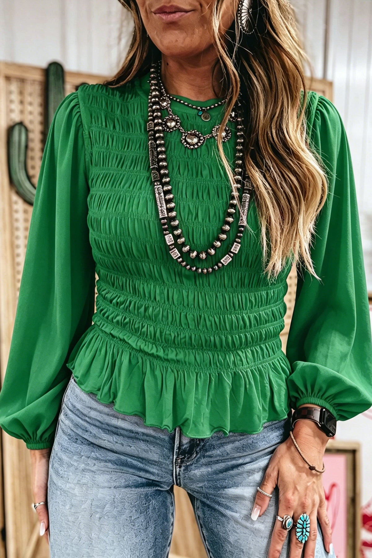A woman wearing a vibrant green Chic Smocked Ruffle Hem Boho Blouse with a smocked bodice and peplum hem, paired with blue jeans and layered silver-toned statement necklaces.