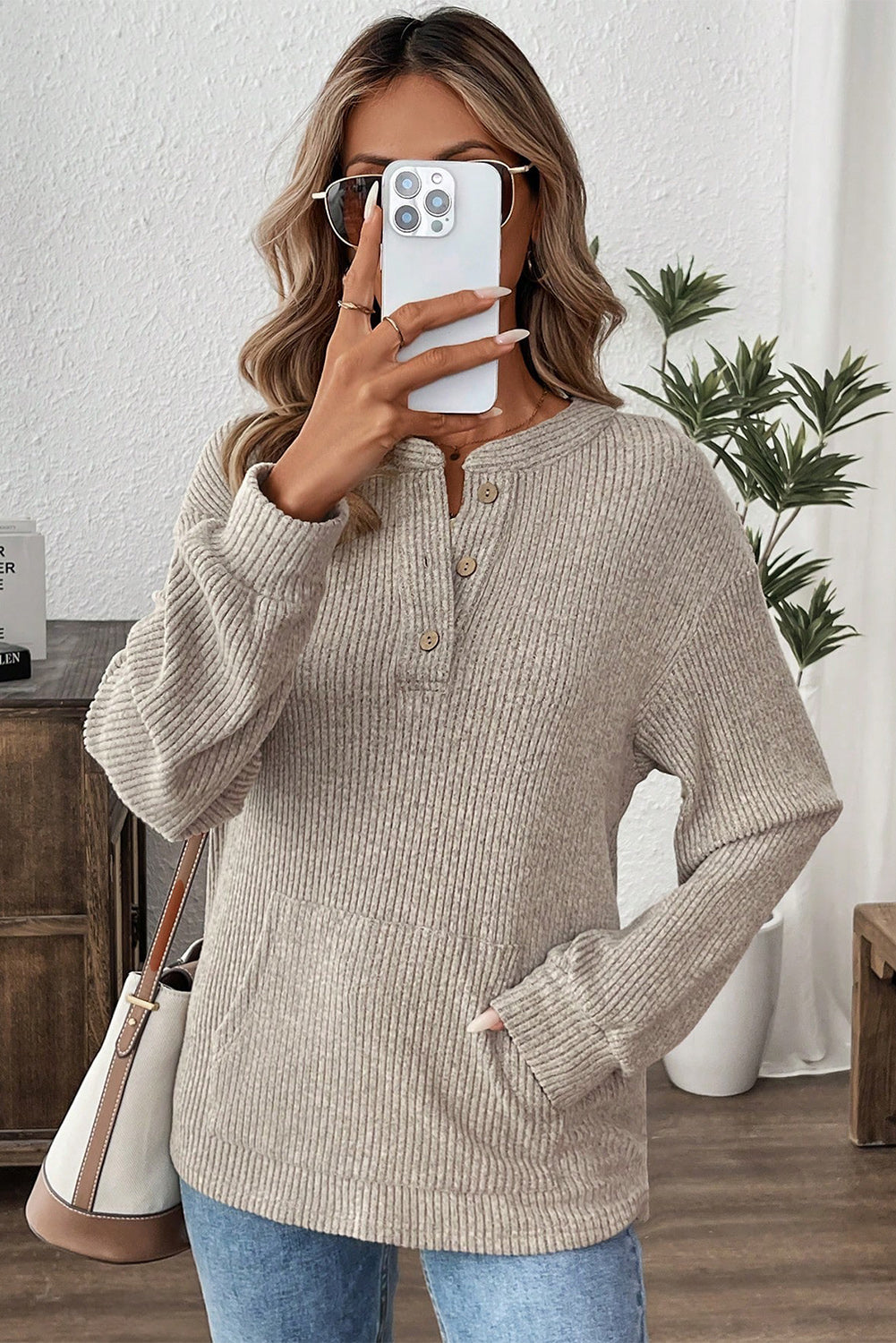 Woman taking a selfie wearing a beige ribbed sweater and blue jeans in a home setting.
