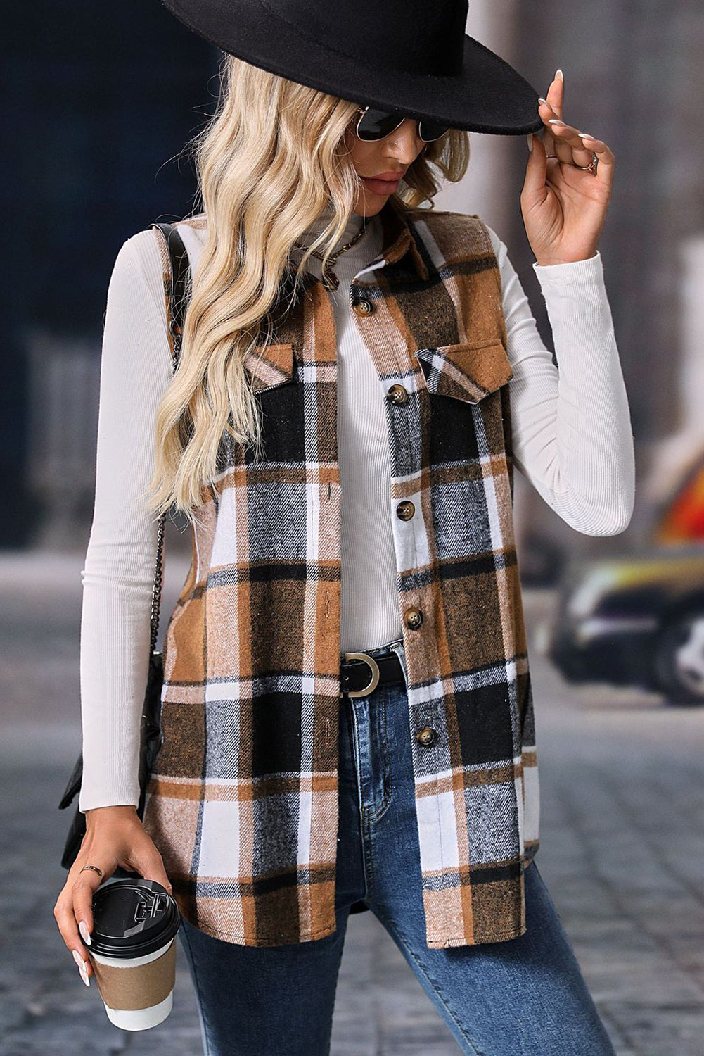 Woman wearing a plaid vest and black hat, holding a coffee cup on a street.
