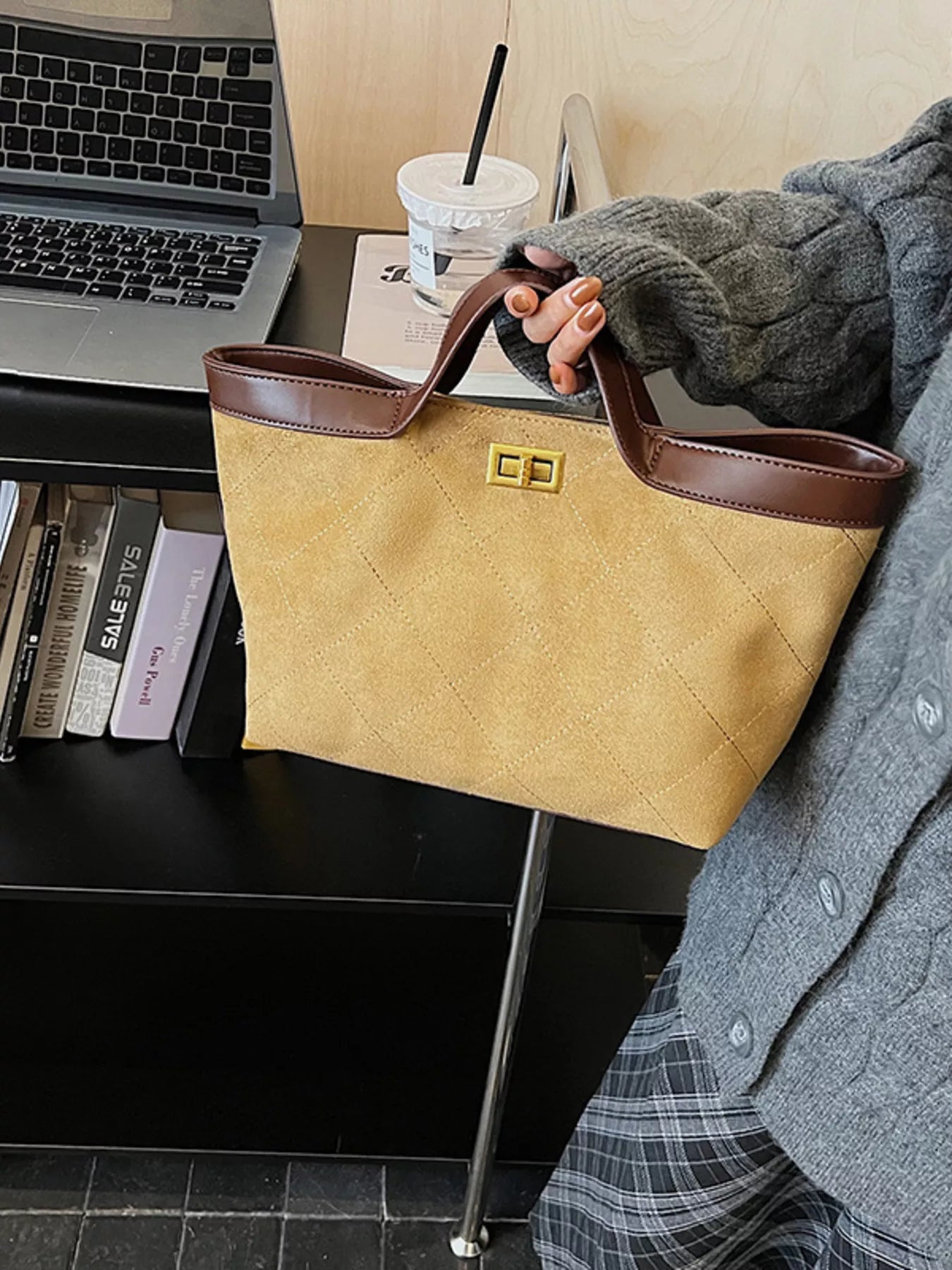 Beige handbag with brown handles held by a person sitting at a desk with a laptop and drink.