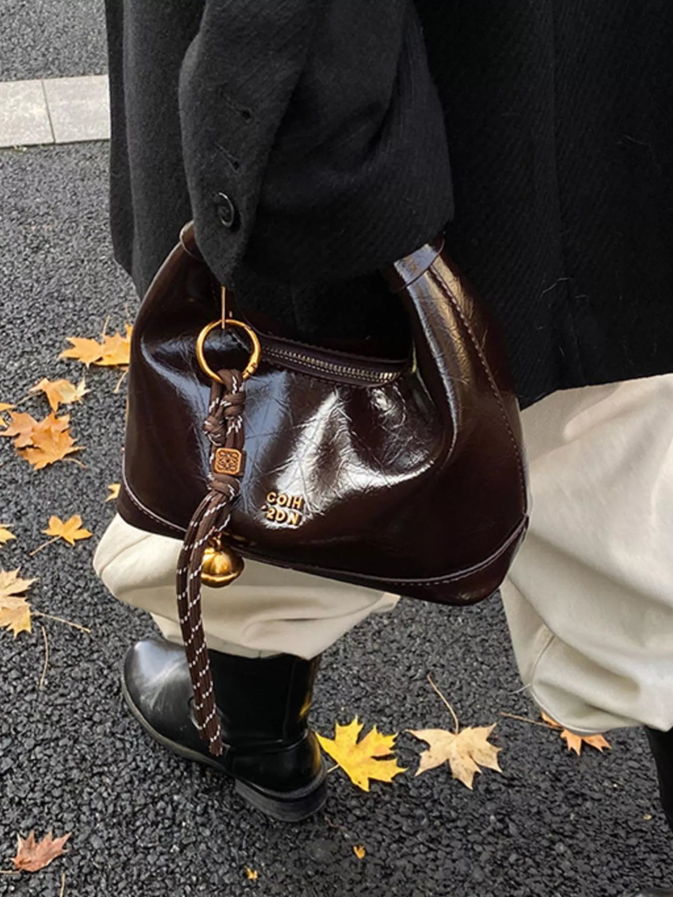 Brown leather handbag with gold accents held by a person on a street with fallen leaves.