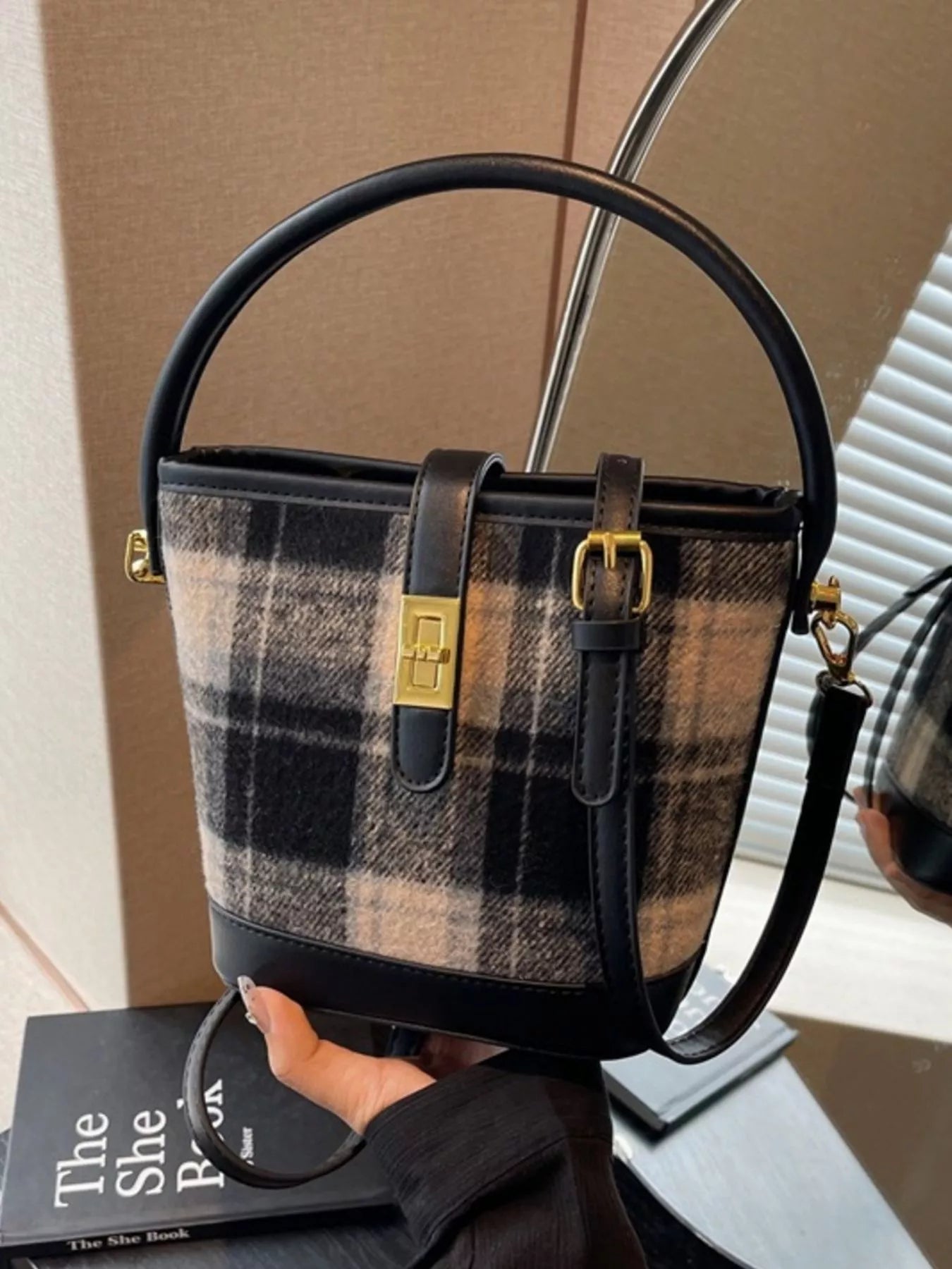 Plaid handbag with black accents held by a person, with a book and chair in the background.