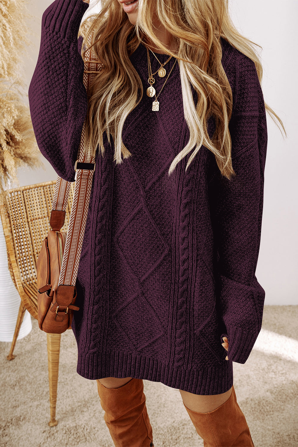 A woman wearing a chunky cable-knit sweater dress in deep plum purple, styled with a brown crossbody bag and brown suede boots 