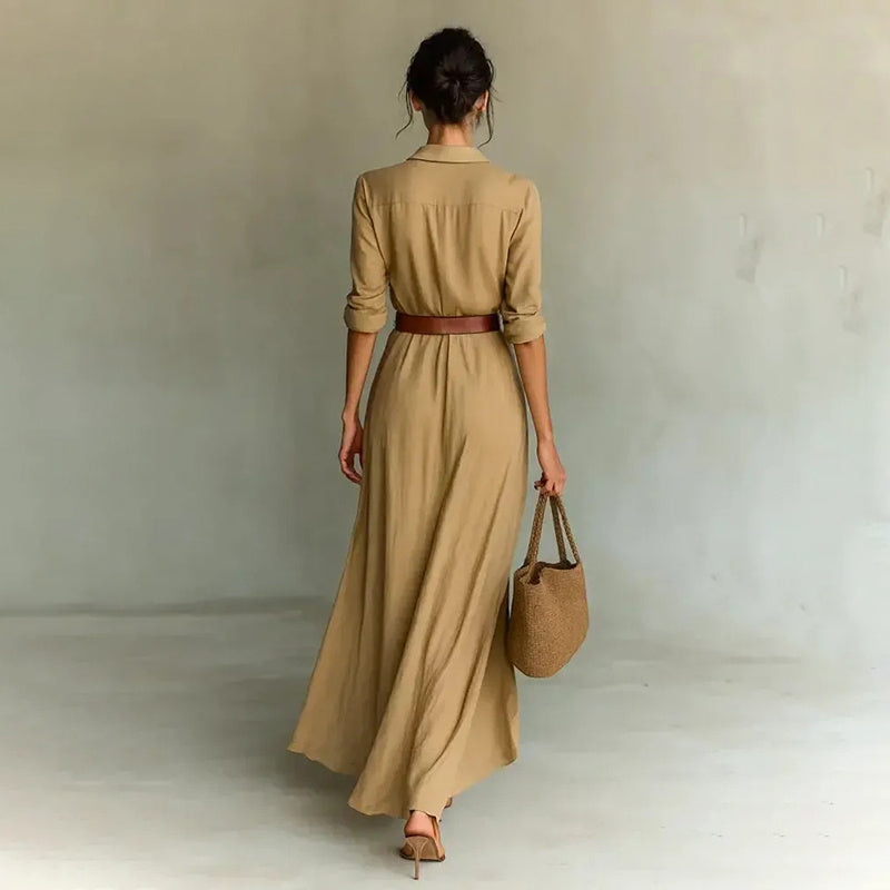 Woman wearing a long beige dress with a belt and holding a matching bag against a plain background